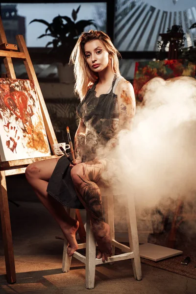 Femme tatouée avec tablier et pinceaux en studio atmosphérique — Photo