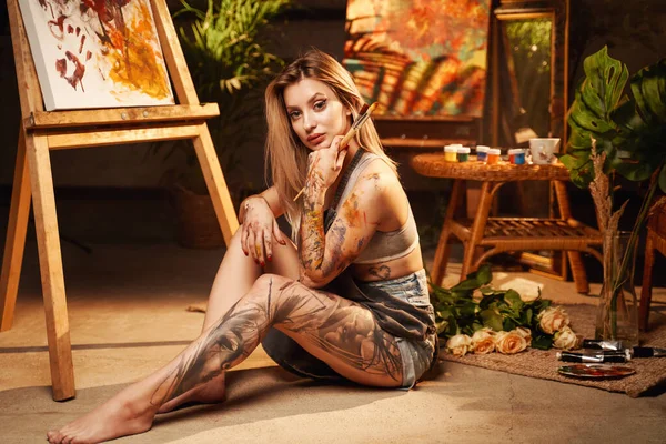 Allegro pittore femminile seduto sul pavimento e che tiene i pennelli in studio — Foto Stock