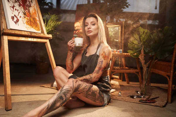 Verführerische grimmige Blondine mit Kaffeetasse in dunkler Kunstgalerie — Stockfoto