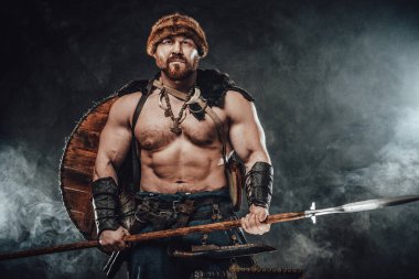 Mızraklı üstsüz Viking dumanlı arka planda poz veriyor.