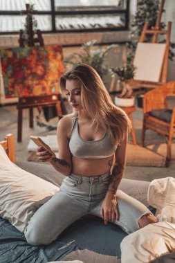 Atölyedeki sarı saçlı kadın telefonunu yatakta kullanıyor.