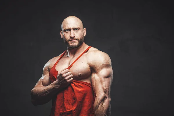 Caucasian bodybuilder poses in dark background pulling his red shirt — Stock Photo, Image