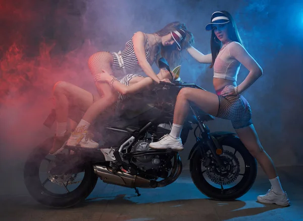 Drie stijlvolle meisjes poseren met custom bike in smokey studio — Stockfoto