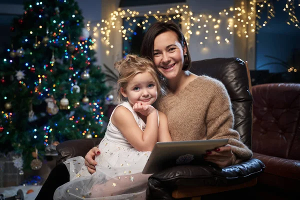 Neşeli anne ve kızı tabletle koltukta oturuyorlar.