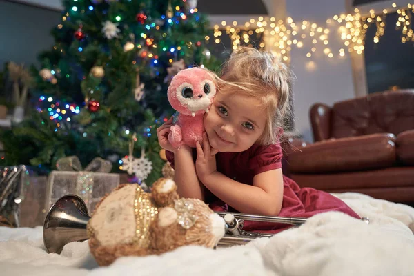 Радостная маленькая девочка с плюшевой игрушкой в уютной комнате с рождественской елкой — стоковое фото
