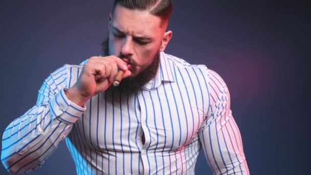 Brutal e bonito fisiculturista com barba e bigode fumar charuto em fundo azul escuro — Vídeo de Stock