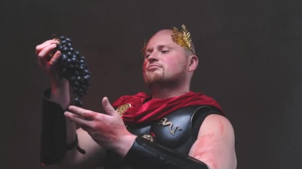 Elite en haarloze Romeinse generaal met krans poseert met druif in donkere achtergrond — Stockvideo