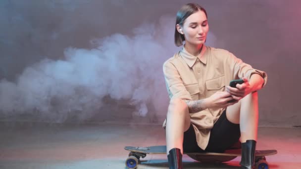 Menina hipster alegre com skate e fones de ouvido posando em estúdio — Vídeo de Stock