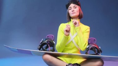 Sarı spor kıyafetleri içindeki oyuncu kız snowboard pozları veriyor.