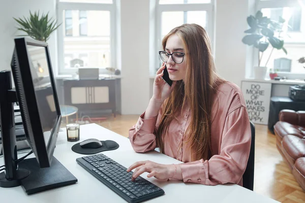 Vrouwelijke kantoormedewerker gebruikt haar telefoon zittend aan tafel met computer — Stockfoto