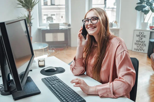 Mulher alegre usa seu telefone sentado à mesa com computador — Fotografia de Stock