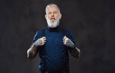 Tişörtlü, kendine güvenen ve şık yaşlı bir adam koyu arkaplanda poz verir.