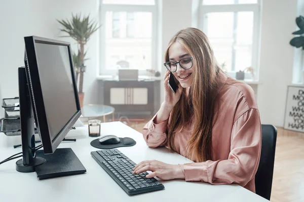 Mulher alegre usa seu telefone sentado à mesa com computador — Fotografia de Stock