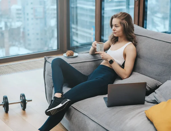 Wesoła kobieta w sportowej odzieży siedzi na kanapie pijąc kawę — Zdjęcie stockowe