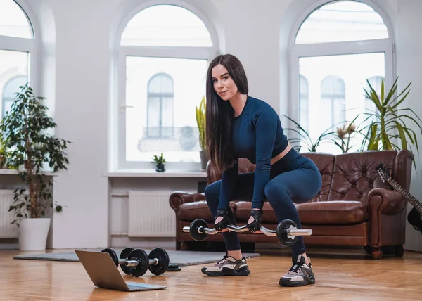 Female athlete works out with barbell using laptop in living room