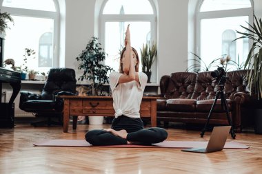 Yoga yapan genç bir kadın gündüz vakti evinde yerde oturuyor.