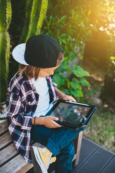 Rapaz bonito com um boné joga um jogo em um tablet sentado em um banco — Fotografia de Stock