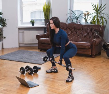 Kadın sporcu oturma odasında dizüstü bilgisayar kullanarak halterle egzersiz yapıyor.