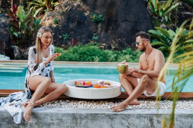 Evli turistler yüzme havuzunun arka planında Asya yemeği yiyip içiyorlar.