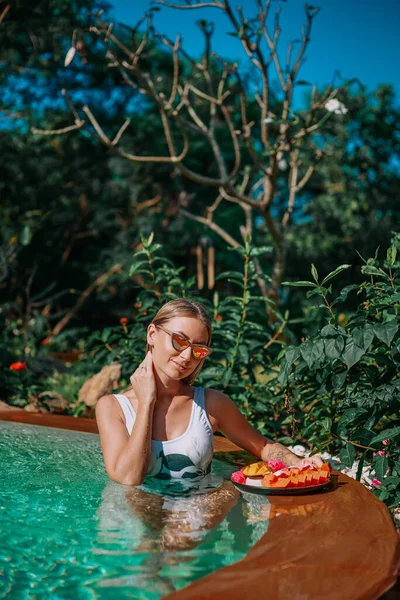 Femme séduisante pose dans la piscine avec de la nourriture tropicale — Photo