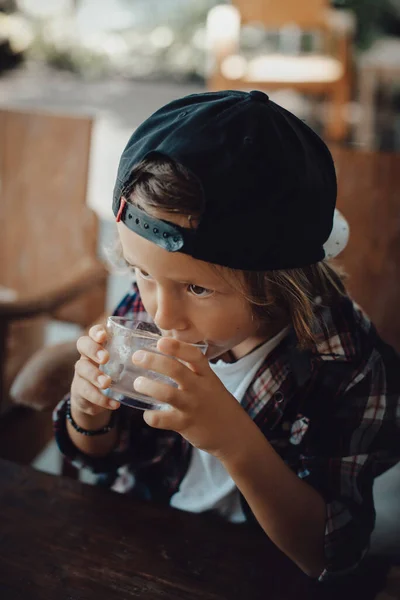 Little boy in shirt with a cap drinks water at table in cafe — Stock Photo, Image