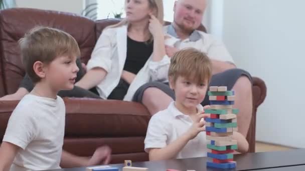 Dois irmãos brincam com blocos de madeira na mesa, pais assistindo alegremente — Vídeo de Stock