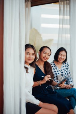 Üç genç Asyalı kadın kameraya bakar ve kapının önünde otururken gülümser.