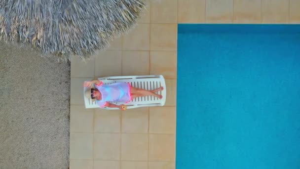 Дронний знімок молодої жінки, що лежить і п'є коктейль біля басейну — стокове відео
