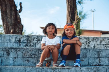 Şapkalı neşeli çocuk ve kaykayın üstünde oturan neşeli kız kardeşi.