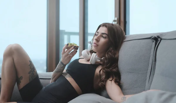 Blanke vrouw met tatoeages en sandwich zit op de bank in appartement — Stockfoto
