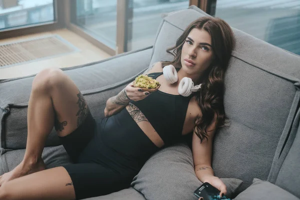 Stijlvolle sportvrouw met tattoos en sandwich liggend op de bank in de kamer — Stockfoto