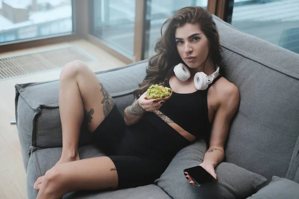 Serieus vrouwelijk model in zwarte sportkleding met sandwich op de bank — Stockfoto