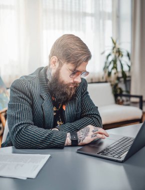 Sakallı stil sahibi serbest yazar gündüz evde dizüstü bilgisayarla çalışıyor.