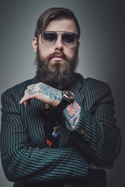 Sakallı ve güneş gözlüklü havalı adam stüdyoda kollarını kavuşturmuş poz veriyor.