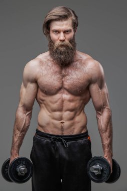 Kas yapısı olan kaba adam gri arka planda dambıllarla poz veriyor.