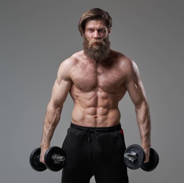 Kas yapısı olan kaba adam gri arka planda dambıllarla poz veriyor.