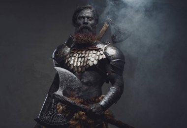 Kaslı mitolojik asker elinde baltayla sisli kameraya bakıyor.