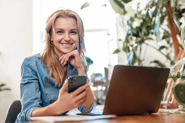 Mulher de cabelos loiros feliz sentada à mesa com laptop e celular — Fotografia de Stock