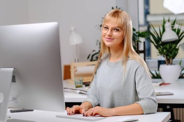 Ofiste neşeli iş kadını gözlükleri ve masaüstü bilgisayarı var.