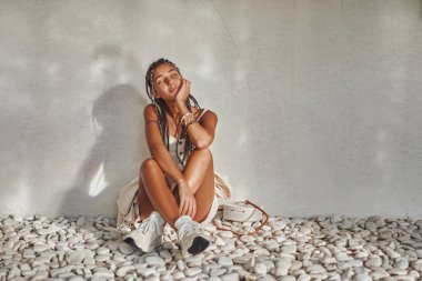 Beyaz yaz elbiseli kadın yaslanmış duvarda oturuyor.