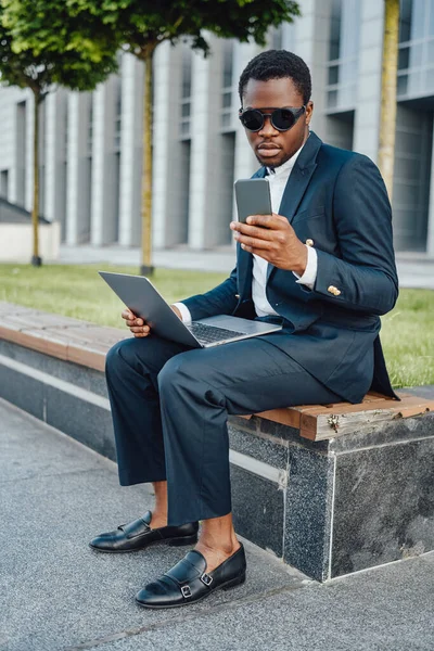 Trabajador corporativo negro serio con portátil mirando el teléfono —  Fotos de Stock