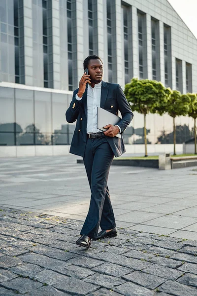 Empresario africano sosteniendo portátil y hablando de teléfono —  Fotos de Stock
