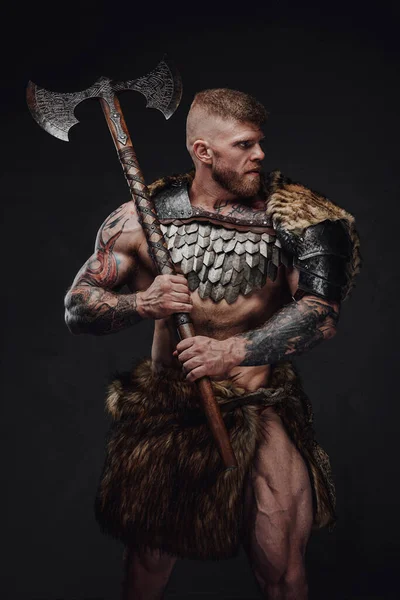 Kaba dövmeli, açık renk zırhlı ve kürklü bir savaşçı karanlık stüdyoda iki eliyle balta tutuyor. — Stok fotoğraf