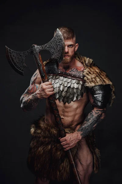 Brutal tattooed warrior wearing light armour and fur holding two-handed axe in dark studio — Stock Photo, Image