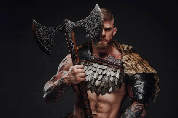 Brutal tattooed warrior wearing light armour and fur holding two-handed axe in dark studio — Stock Photo, Image