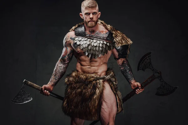 Guerreiro tatuado brutal usando armaduras leves e machados de pele no estúdio escuro — Fotografia de Stock