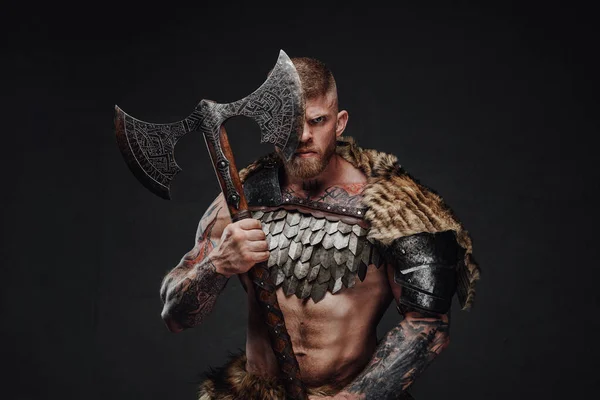 Brutal tattooed warrior wearing light armour and fur holding two-handed axe in dark studio — Stock Photo, Image