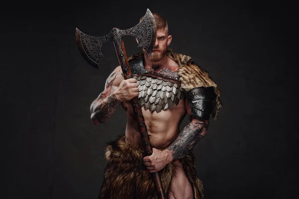 Brutal tatuado guerreiro vestindo armadura leve e pele segurando machado de duas mãos no estúdio escuro — Fotografia de Stock