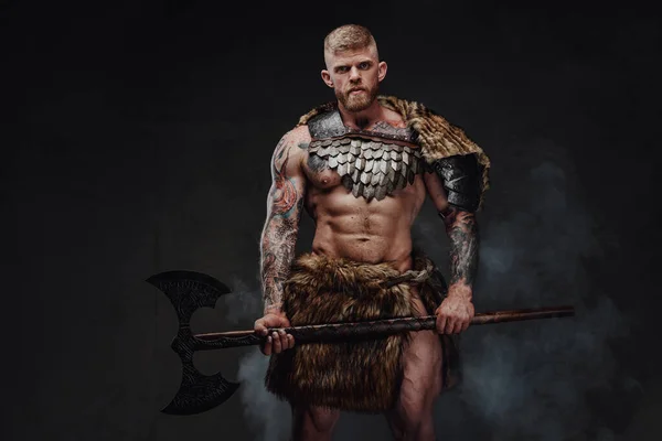 Brutal tattooed warrior wearing light armour and fur holding two-handed axe in dark studio — Stock Photo, Image