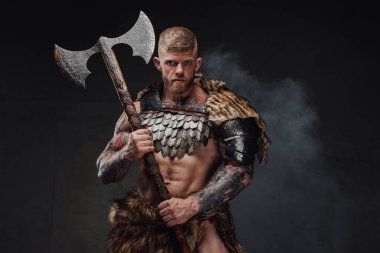 Kaba dövmeli, açık renk zırhlı ve kürklü bir savaşçı karanlık stüdyoda iki eliyle balta tutuyor.